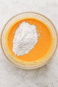 A clear glass bowl containing an orange batter with a heap of white flour added on top, placed on a marble surface.