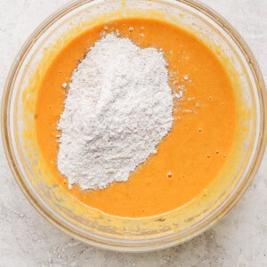 A clear glass bowl containing an orange batter with a heap of white flour added on top, placed on a marble surface.