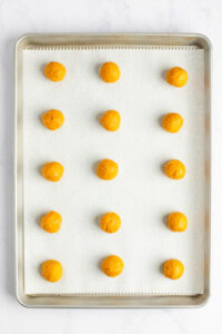 A baking tray with 15 evenly spaced, round, orange dough balls placed on a parchment-lined surface.