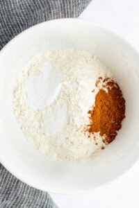 A white bowl containing flour, cinnamon, and baking powder, with a striped cloth partially visible in the background.