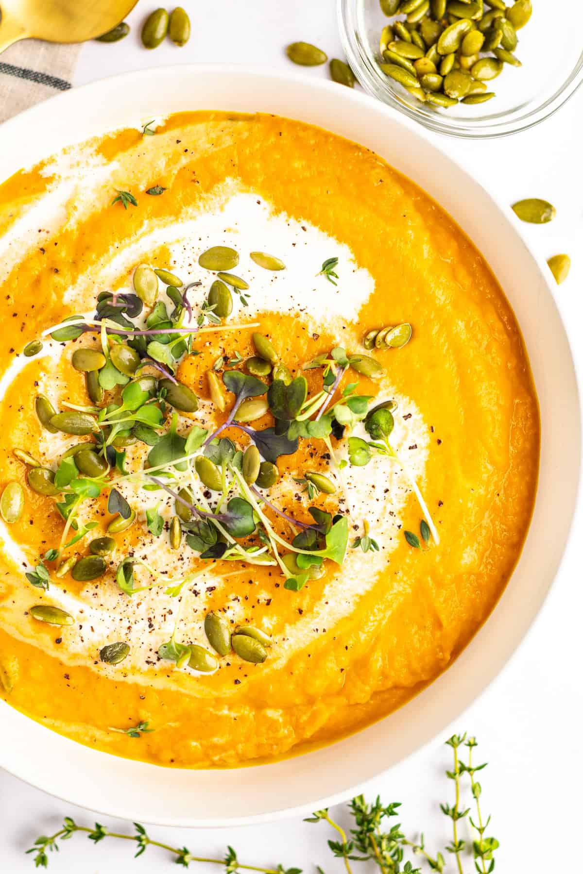 A bowl of creamy pumpkin soup garnished with a swirl of cream, microgreens, and pumpkin seeds, with additional pumpkin seeds and thyme sprigs on the side.