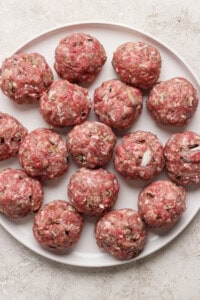 A plate filled with raw, evenly-sized meatballs, arranged in a circular pattern on a white surface.