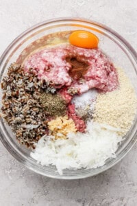 A glass bowl containing ground meat, an egg yolk, wild rice, spices, breadcrumbs, minced garlic, and shredded onions.