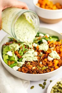 A hand pouring green herb dressing from a jar onto a bowl containing quinoa, roasted vegetables, greens, seeds, and crumbled cheese with another bowl of roasted vegetables in the background.