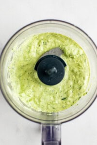 A bowl of green and white sauce sits inside a food processor with a blade in the center.