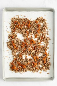 A baking tray covered with parchment paper holds a single layer of evenly spread, mixed quinoa with a toasted appearance.