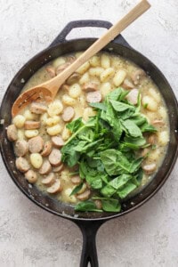 A skillet containing gnocchi, sliced sausages, and fresh spinach, with a wooden spoon resting inside.