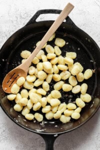 A cast iron skillet filled with cooked gnocchi, stirred with a wooden spoon, on a light gray textured surface.