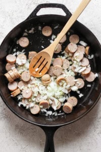 A black skillet containing sliced sausages and diced onions being stirred with a wooden spoon.