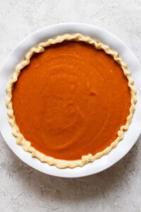 A round, unbaked pumpkin pie with a crimped edge in a white pie dish sits on a light-colored textured surface.