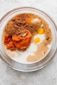 A glass bowl containing pumpkin puree, two eggs, milk, and various dry spices, ready to be mixed.