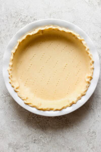 A pie crust in a white pie dish with a crimped edge and multiple small fork holes in the base, placed on a light textured surface.