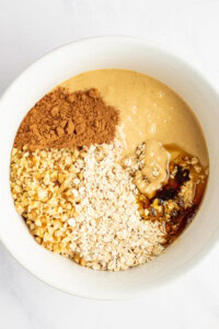 A white bowl containing peanut butter, chopped nuts, oats, cocoa powder, and honey.