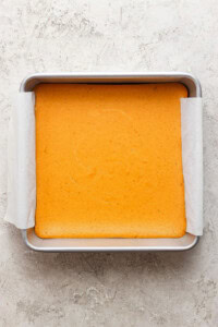 A square pan with a light golden-brown baked cake lined with parchment paper on a textured, light-colored surface.