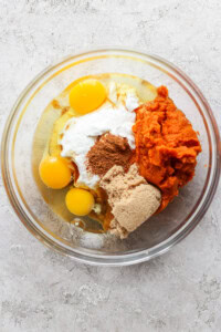 A glass mixing bowl containing raw eggs, sour cream, pumpkin puree, brown sugar, and spices, ready to be mixed.