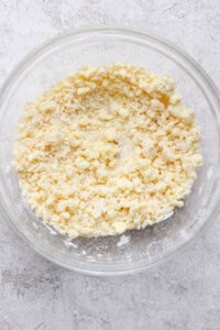 A clear glass bowl containing a crumbly mixture of flour and butter, sitting on a textured gray surface.