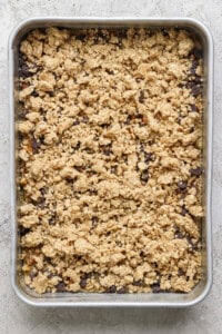 A rectangular baking tray filled with an unbaked dessert mixture featuring a crumbly oat and nut topping, placed on a textured light-colored surface.
