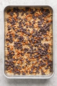 A silver baking pan containing an uncut dessert topped with chocolate chips and chopped nuts.