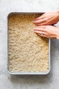 Hands press down a mixture in a square baking pan. The mixture contains visible oats and is evenly spread across the pan.