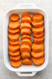 A white baking dish filled with sliced sweet potatoes arranged in three rows on a light surface.