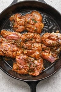 Raw marinated chicken thighs in a dark cast iron skillet, ready to be cooked. The marinade includes herbs and a reddish sauce. The skillet rests on a light, textured surface.