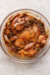 A clear glass bowl containing marinated pieces of raw chicken mixed with herbs and spices in a thick, reddish-brown sauce.