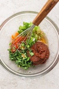 A glass bowl containing chopped green herbs, red sauce, a thick brown paste, and a whisk with a wooden handle on a light-colored surface.