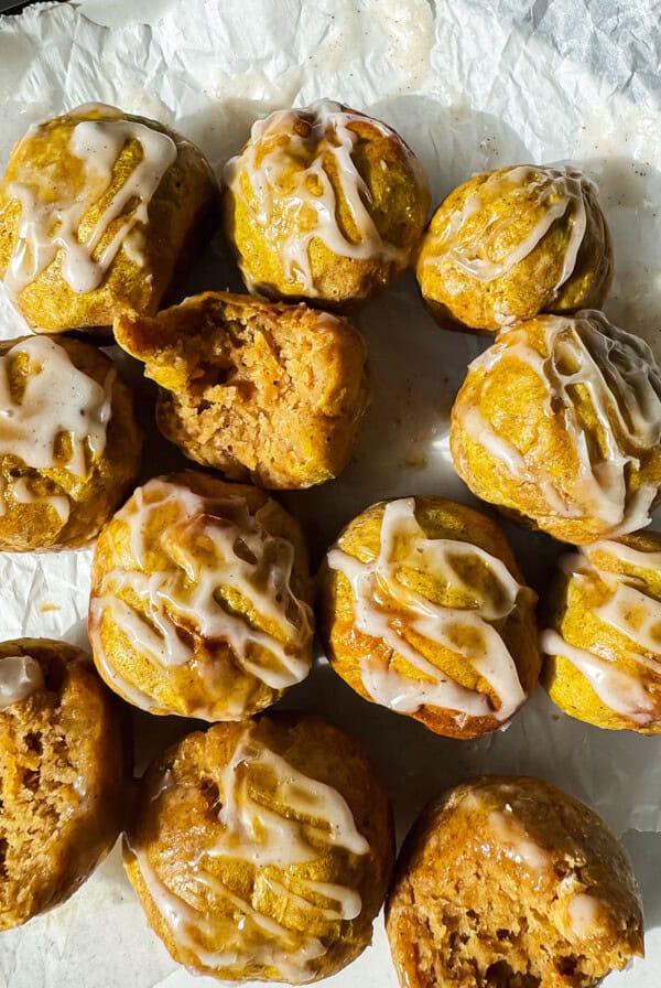 A dozen small, round cakes with white icing drizzle, some with bites taken out, are arranged on crumpled parchment paper under natural light.