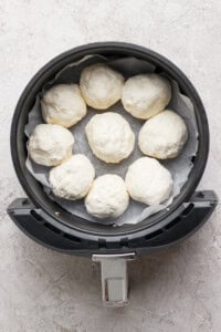 An air fryer basket lined with parchment paper holds eight uncooked dough balls in a circular arrangement.