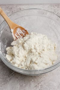 A wooden spoon rests in a glass bowl filled with a mixture of flour and a white ingredient, likely dough, on a textured countertop.