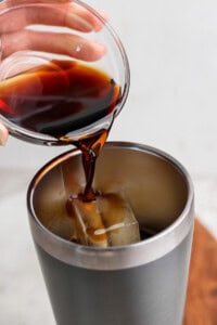 Pouring dark liquid into a stainless steel tumbler with ice.