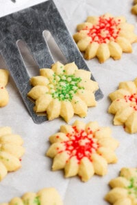 A spatula lifts a star-shaped, green-sprinkled cookie. Other cookies with red sprinkles are on a parchment-lined surface.