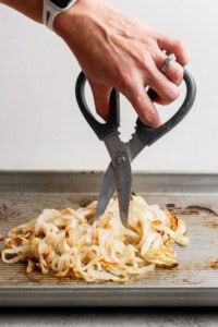 A hand using kitchen scissors to cut cooked onions on a baking sheet.