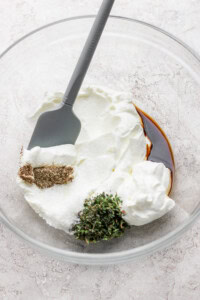 A mixing bowl with yogurt, soy sauce, herbs, and spices, stirred with a gray spatula.